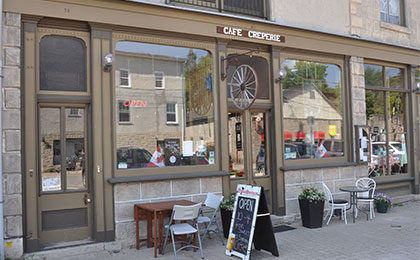 Store front for Cafe Creperie with two large windows and a window-panelled door into a limestone building, with cafe tables and chairs and a sandwich board out front.
