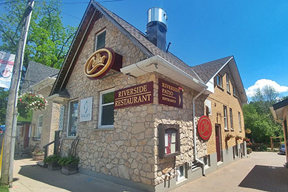 The Cellar restaurant storefront faces the sidewalk of East Mill Street in Elora with a stone exterior that turns to brick in the back portion of the building. A large oval sign sits centre of the upper level of the building with a burgundy background and the business name Cellar in gold letters. Another sign using the same colours on the north corner of the building reads: riverside restaurant. On the upper west corner of the building another sign with an arrow directs patrons to the riverside patio behind the building. Below it, a large wood case features the menu. Walking down the side of the building along an interlocking stone pathway, a large oval sign attached to the side of the building reads Cellar. Many windows and a side door are in the more modern brick extension of the building that leads to an outdoor patio overlooking the river.