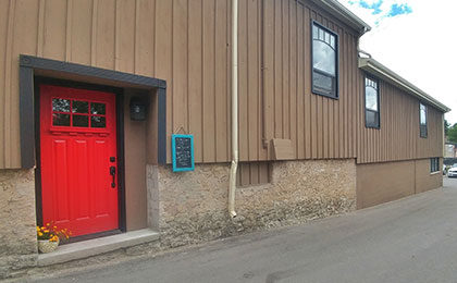 Exterior of a brown siding building with stone foundation and a large, bright red door with black trim. A flower pot sits outside the door entrance. A small blue framed chalkboard details the daily specials.