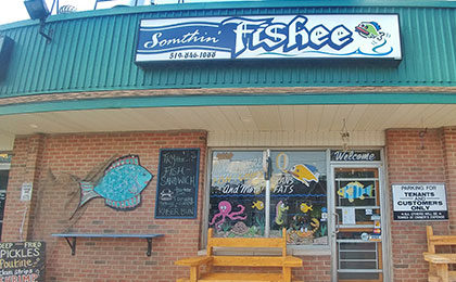 Exterior brick plaza storefront, with a window and door decorated in colourful cartoon fish and ocean creature paintings. Outside the store, a large blue fish plague rests on the wall next to a black chalkboard with the daily specials, and two wooden seating benches adorn each side of the front glass door.