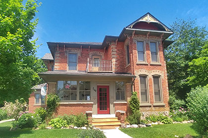 The Flying Leap Bed & Breakfast is a large two-storey Victorian home on a corner lot in downtown Elora. A heritage red brick home with ornate brick patterns, the house features many windows and a small balcony on the second floor. It has a large front closed-in porch accessible by four steps that lead to a large red front door. There are gardens trimmed with stone and a green lawn out with a concrete path leading guests into the house.