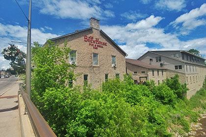 The historic limestone two-storey building of the Brew House Pub & Grill overlooks the Grand River as it winds through downtown Fergus. Here there are many trees and bushes along the rocky shoreline leading up to the wall. Four windows overlook the river, while above three windows offer a view to B & B guests. The restaurant on the lower level includes a patio at the back of the building, connecting it to a series of other heritage stone buildings with modern roofs and additions.