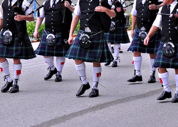 A tartaned clad bagpipe band marches along a road. The image doesn't show the heads of the all-male participants. It shows the bagpipers from the shoulders down, wearing black vests with silver square buttons. The tartan plan kilts are dark blue with green, purple and white patterns. Each piper wears a silver and black leather sporan, white knit socks up to the knee with red sashes and black leather lace-up kilt shoes. This photo is of members of the Fergus Pipe Band.