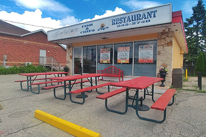 An exterior image of a building with stone exterior in the front, with four large window panels and a glass door at the far left to enter the establishment. Out front are 3 red picnic tables bordering the parking lot. A red bench backs up against the windows. Along the extension of the roof is a sign that reads Golden Fish & Chips Restaurant.