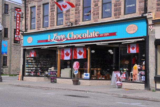 An outside image of the storefront for I Love Chocolate in downtown Fergus. The store is located on the street level of a stone block heritage building. The storefront is all windows that are flat with the building and then turn into it to create two separate alcoves, with two doors that are in the alcoves. Across the length of the store, above the windows, is a bright blue wooden panelled wood sign that bears the store logo: I Love Chocolate and Gifts. On each end of those words is a an oval sign in light brown. On the left of the sign it reads: I love that chocolate. On the right of the main sign, the oval sign reads: I love that gift. The windows offer a full view of the interior of the store. There are two sandwich boards outside each end of the building displaying their fudge and daily specials.