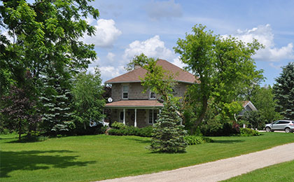 Maplecrest Farm (front) Limestone two-story farmhouse with a large porch and a green lawn with big trees, a gravel driveway.