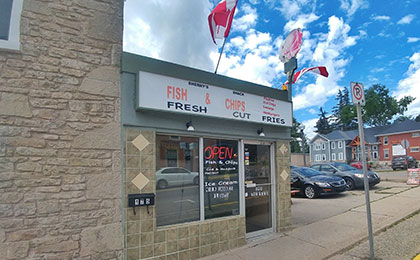 Exterior of a small brick building with double large window panes and a glass door. Above the window and doors is green siding with a large white sign that reads: Fish and Chips, Fresh Cut Fries. Three Canadian flags are attached to the top of the sign. A pedestrian sidewalk is out front of the store.