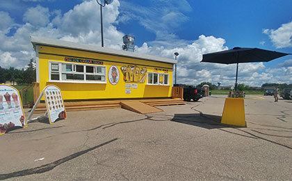 A small bright yellow chip wagon building with two large order windows on each end of the front of the building, with the Fry Shack's cartoon French fry logo between them. Stairs and an accessibility ramp lead up to the building.