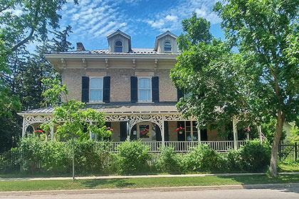 A historic home with a fully renovated carriage house has become the Drew House Bed & Breakfast in Elora. The main house, a Victorian-style stone house with two-storeys and an attic with dormers features a wrap around porch with ornate details along the roof and railings in a light paint colour. The grand home has three windows on the second storey with black shutters. Under the patio is a front door, centered between two windows, all with long black shutters. The veranda has hanging baskets of flowers and a garden of bushes and trees that obscure the foundation of the house.