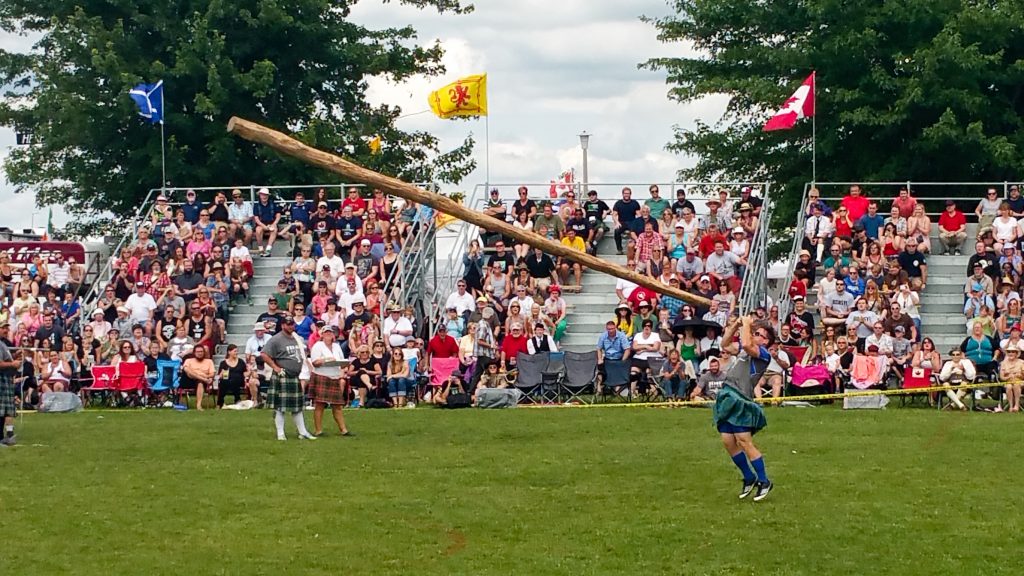 Fergus Scottish Festival and Highland Games caber toss event with an athlete throwing the caber in mid-air before a crowd of onlookers on the bleachers.